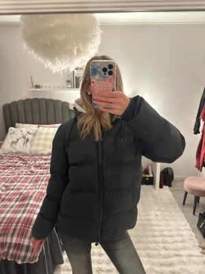 Mörkblå pufferjacka från Rains - Mörkblå pufferjacka från Rains i storlek S. Jackan har huva, dragkedja framtill och är tillverkad i syntetmaterial som är både vattentätt och vindtätt. Den är isolerad och har en clean, oversized look med långa ärmar och praktiska fickor.