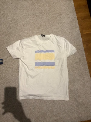 Vit t-shirt från A.P.C. med tryck - Vit t-shirt från A.P.C. i bomull med korta ärmar och coolt grafiskt tryck i ljusgul och ljuslila på bröstet. Klassisk passform och rund halsringning. Perfekt för en clean och stilren look.