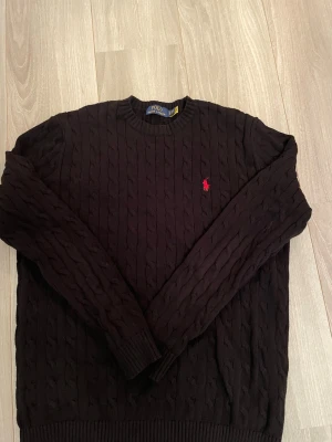 Svart kabelstickad tröja Polo Ralph Lauren - Svart kabelstickad tröja från Polo Ralph Lauren med klassisk rund hals och röd broderad logga på bröstet. Tröjan har långärmade ribbade muddar och är gjord i mjuk bomull. Perfekt för dig som gillar stilrena och tidlösa plagg.
