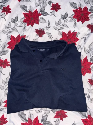 Mörkblå piké från Race Marine - Säljer en stilren mörkblå pikétröja från Race Marine med klassisk krage och två knappar framtill. Pikén har korta ärmar och ett diskret RM-broderi på bröstet. Perfekt för dig som gillar enkel och snygg design.