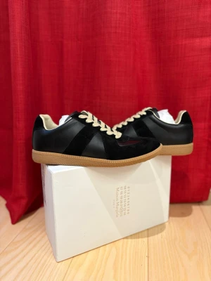 Maison Margiela Gats Skor - Maison Margiela sneakers i svart skinn och mocka med beige snörning och innersida. Sulan är i gummi med klassiskt mönster och logotyp under foten. Snygg kontrast mellan svart ovandel och ljusbrun sula, perfekt för dig som gillar stilrena detaljer. Allt tillkommer