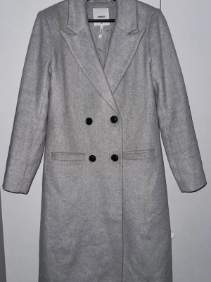 Grå kappa från Object, strl 34 - Säljer en stilren grå kappa från Object i storlek 34. Dubbelknäppt framtill med svarta knappar och klassisk krage. Långärmad modell med knappar vid ärmslut och raka fickor. Perfekt för dig som gillar minimalistisk och clean look.