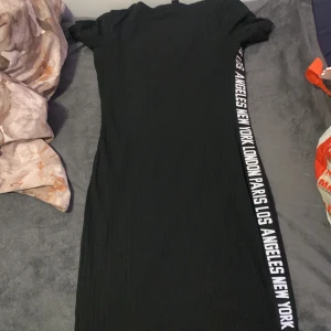 Svart ribbad klänning från SHEIN - Svart ribbad kortklänning från SHEIN med korta ärmar och cool textdetalj längs ena sidan med namn på olika storstäder. Tight passform och stretchigt material som sitter snyggt på kroppen. Perfekt för dig som gillar streetstyle och enkel design.