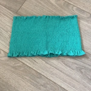 Turkos bandeau tubtopp - Säljer en turkos bandeau tubtopp med smockad struktur och volangkant upptill och nedtill. Perfekt för dig som gillar färgglada och trendiga toppar. Enkel att matcha med jeans eller kjol för en cool look.