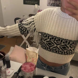 Vit och svart croppad stickad tröja - Säljer en croppad stickad tröja från H&M i vit med svart grafiskt mönster och fransar längs ärmarna. Tröjan har ribbad struktur, rund hals och långa ärmar. Perfekt för dig som gillar statement-plagg med coola detaljer. Rensar garderoben, och kan därför gå ner i pris vid snabb affär!
