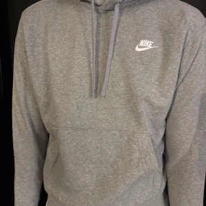 Grå hoodie från Nike med huva - Grå hoodie från Nike med klassisk logga broderad på bröstet. Tröjan har huva med snörning, stor magficka och är tillverkad i mjuk bomullsmix med fleece på insidan. Perfekt för chill eller träning, och har långa ärmar för extra komfort.