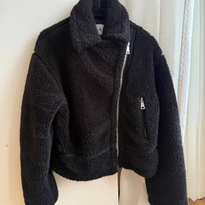 Svart teddyjacka från NA-KD - Mysig svart teddyjacka från NA-KD med stor krage och sned dragkedja framtill. Jackan har långärmade ärmar och en cool, lite boxig passform. Perfekt för att hålla värmen. Fleece-liknande material och snygga detaljer med dragkedjor.