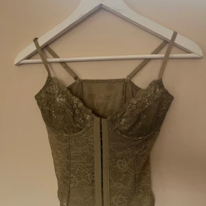 Beige spetskorsett med snörning - Säljer en beige korsett med tunna axelband och blommig spets över hela toppen. Framsidan har snygg snörning och kuporna är lätt formade för extra stöd. Perfekt för dig som vill ha en feminin och trendig look med transparent känsla. Använd bara en gång