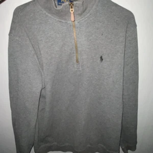 Grå half zip tröja från Polo Ralph Lauren - Säljer en stilren grå tröja från Polo Ralph Lauren med half zip-dragkedja och klassisk broderad logga på bröstet. Tröjan har lång ärm och är tillverkad i mjuk bomull, perfekt för lager-på-lager under kyligare dagar.