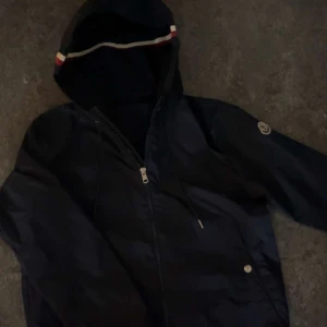 Mörkblå  vindjacka från Moncler pris kan diskuteras  - Mörkblå vindjacka från Moncler med huva och dragkedja framtill. Jackan har en snygg detalj med röd, vit och blå rand på huvan samt Moncler-logga på ärmen. Materialet är lätt och känns sportigt, perfekt för dig som gillar stilren design.