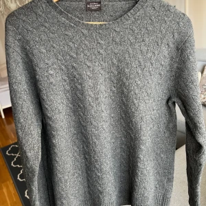 Rose & Born soft blend Kashmir tröja - Snygg grå kabelstickad tröja från Rose & Born i mjuk ullblandning (70% ull). Rund halsringning och ribbade muddar vid ärmslut och nederkant. Perfekt för kyliga dagar och enkel att matcha med jeans eller chinos. Väldigt gott skick, de är inga noppor utan det är ullen som ska se ut så där. Storlek 50, passar M. 