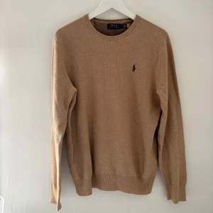 Beige stickad tröja från Polo Ralph Lauren - Snygg beige stickad tröja från Polo Ralph Lauren med klassisk rund hals och diskret logga broderad på bröstet. Tröjan har långa ärmar och ribbade muddar vid ärmslut och nederkant. Perfekt för dig som gillar stilrena och tidlösa plagg.