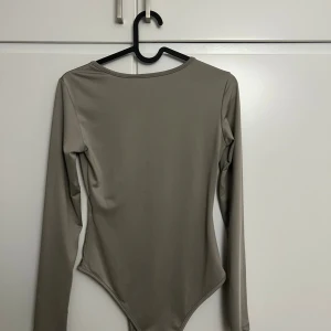 Beige långärmad bodysuit - Säljer en stilren beige/grå bodysuit med lång ärm och rund hals. Toppen har en tight passform och är tillverkad i ett mjukt, stretchigt syntetmaterial som sitter snyggt mot kroppen. Perfekt att styla med jeans eller kjol för en clean look.