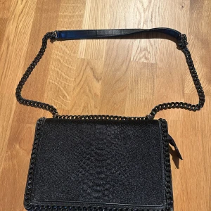 Svart axelväska med kedja - Snygg svart axelväska med kedjerem och detaljerad framsida med reptilmönster. Väskan har flera fack, dragkedja på baksidan och röd insida som ger en cool kontrast. Perfekt storlek för det viktigaste och stilren look.