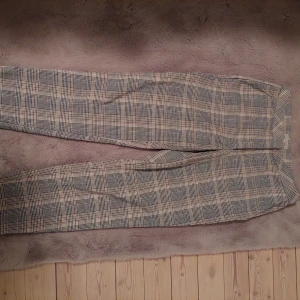 Rutig kostymbyxa från H&M, stl 38 - Snygga kostymbyxor från H&M i klassiskt rutigt mönster med toner av beige, svart och grått. Byxorna har rak passform och normal midja, perfekta för en stilren look. Materialet är mjukt och bekvämt, passar till många olika outfits.