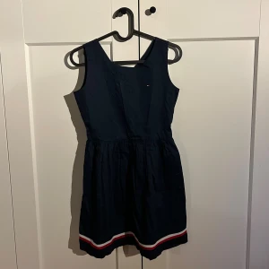 Mörkblå Tommy Hilfiger klänning  - Supersnygg mörkblå ärmlös klänning från Tommy Hilfiger med u-ringning och dragkedja i ryggen. Kjolen har en cool röd, vit och blå rand längst ner och ett litet märke på bröstet. Klänningen är väldigt stilren och bekväm. Perfekt för både kalas, middagar och att ha hemma. 