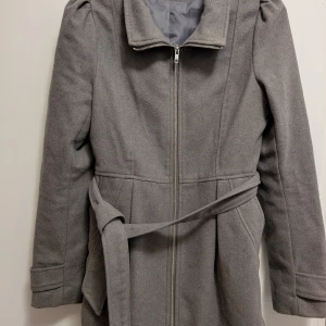 Grå kappa med bälte och dragkedja - Stilren grå kappa med dragkedja framtill och matchande bälte i midjan. Jackan har klassisk krage, långa ärmar och en rak, något figursydd passform. Perfekt för kyligare dagar och enkel att matcha med olika outfits.