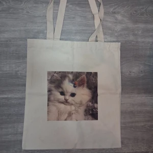 Beige shoppingväska med kattmotiv - En beige shoppingväska i ett tjockt material med tryck av en söt kattunge med hårklämmor på huvudet. Väskan har dubbla axelband och är perfekt för att bära med sig böcker, laptop eller annat du behöver under dagen. Enkel och stilren design med gullig touch. Ca 38x34cm 