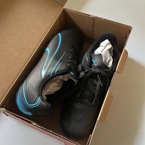 (helt nya) Svarta Puma fotbollsskor med blå detaljer - Säljer ett par svarta fotbollsskor från Puma med snygga blå detaljer och dobbar. Skorna har snörning och är tillverkade i syntetmaterial. Perfekta för dig som vill ha stilrena och sportiga skor på planen. Puma-loggan syns tydligt på sidan och innersulan.
