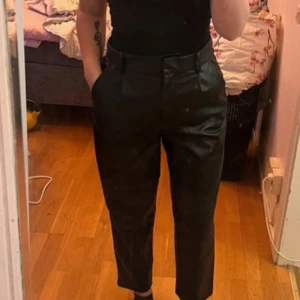 Svarta byxor i skinnimitation - Säljer ett par svarta byxor i skinnimitation med hög midja och raka ben. Byxorna har bälteshällor och sidfickor. Dom är i storlek L och sparsamt använda