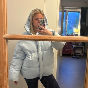 Ljusblå pufferjacka med huva - Säljer en ljusblå pufferjacka med stor huva och dragkedja framtill. Jackan är oversized och har breda kanaler som ger en riktigt fluffig look. Perfekt för kalla dagar när man vill vara både varm och snygg.