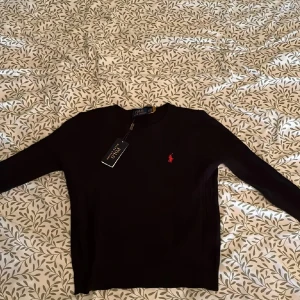 Svart stickad tröja Polo Ralph Lauren - Svart stickad tröja från Polo Ralph Lauren i storlek S. Klassisk rund hals och långärmad modell med diskret röd logobroderi på bröstet. Tröjan har ribbade muddar och ett stilrent, tidlöst utseende som funkar till många olika looks.