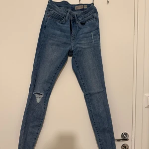 Mid rise slim jeans XS från Vero Moda - Snygga blå jeans från Vero Moda i modellen mid rise slim. Jeansen har en tight passform, klassisk femficksdesign och slitning på ena benet. Tillverkade i mjukt denim med råa kanter nertill för en trendig look.
