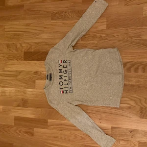 Grå långärmad tröja från Tommy Hilfiger - Snygg grå långärmad tröja från Tommy Hilfiger med stort tryck på bröstet och klassisk logga. 