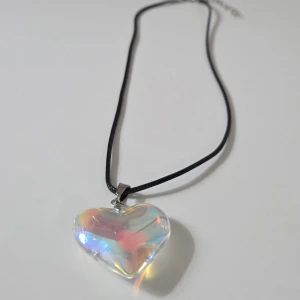 Regnbågshalsband med hjärthänge i glas, 45 + 5 cm - Unikt halsband med ett glittrigt hjärthänge i regnbågsfärgat glas. Hänget har en skimrande effekt med inslag av rosa, blått, gult och lila. Svart snöre och silverfärgad fästanordning ger en snygg kontrast. Perfekt för dig som vill sticka ut med färg och cool design.