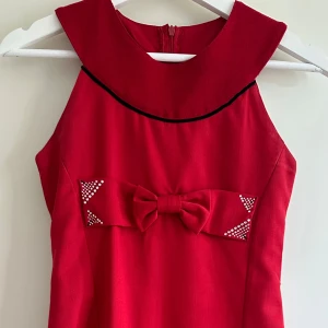 Röd klänning  - Supersöt röd klänning med bred krage, svart kantdetalj och en stor rosett framtill dekorerad med små silvriga nitar. 