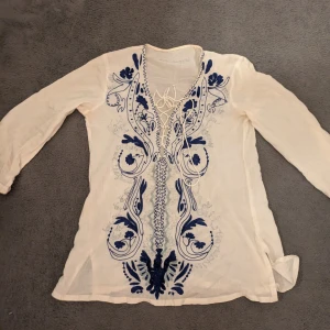 Beige blus med blå broderi - Säljer en beige blus med lång ärm och v-ringad hals med snörning. Blusen har detaljerat blått broderi i ett blommigt mönster framtill som ger en bohemisk vibe. 