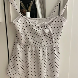 Vit prickig linnetopp från Seed Heritage - Supersöt vit linnetopp med svarta prickar från Seed Heritage. Toppen har smala axelband, knyt framtill och en lätt utställd form som ger en chill vibe. Perfekt för varma dagar och enkel att styla med jeans eller kjol. Materialet känns mjukt och luftigt.