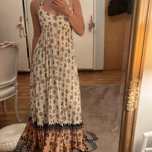 Boho långklänning med paisleymönster - Luftig långklänning i boho-stil med halterneck och paisleymönster. Klänningen är vit med inslag av beige och svart, samt en bred bård i botten med mönster i orange och blått. Perfekt för sommardagar och har en flowy passform.