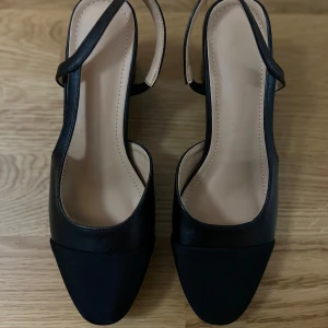 Svarta pumps med klack och öppen häl - Stilrena svarta pumps med öppen häl och bred klack. Skorna har en fyrkantig tå och rem runt hälen. Ovandelen är i svart material med en matt textil framtill och slät finish på sidorna. Perfekta för dig som gillar klassisk och chic stil. Nypris 599kr säljs för 450 kvitto finns oanvända Storek 38,5 