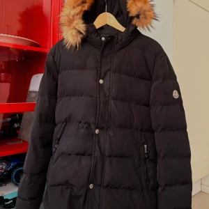 Svart Moncler dunjacka med päls - Svart dunjacka från Moncler med fluffig päls på huvan och quiltad design. Jackan har dragkedjor på fickorna, tryckknappar framtill och Moncler-logga på ärmen. Perfekt för kalla dagar och har en snygg, klassisk look. OBS! Observera skadan på jackan, den måste till skräddaren men annars mycket fin
