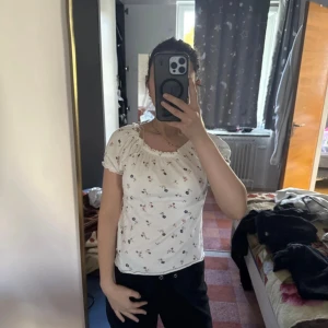 Vit blommig blus med puffärm - Supersöt vit blus med små blommor i rosa och blått. Modellen har korta puffärmar, resår vid halsringning och ärmslut samt lätt rynkade detaljer. Tunt, luftigt tyg som känns perfekt för varmare dagar. Söt och feminin stil som passar till jeans eller kjol.