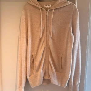 Beige hoodie i kashmir från Soft Goat - Mysig beige hoodie från Soft Goat i lyxig kashmir. Tröjan har dragkedja framtill, huva med snörning och två fickor. Perfekt för dig som vill ha en stilren och skön look med premiumkänsla.