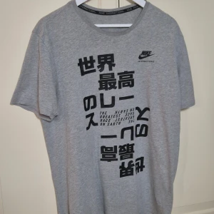Grå Nike t-shirt med tryck L - Snygg grå t-shirt från Nike med svart grafiskt tryck och japanska tecken på framsidan. Modellen har rund hals och korta ärmar. Tillverkad i mjuk 100% bomull för skön känsla. Perfekt för dig som gillar streetwear och sportig stil. I fint skick!