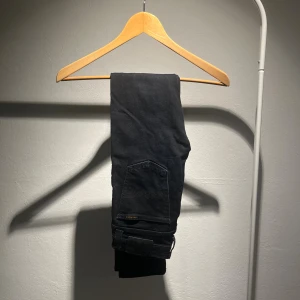 Svarta jeans från Nudie Jeans - Snygga svarta jeans från Nudie Jeans med klassisk femficksdesign och vågig broderad detalj på bakfickan. Jeansen har en mörk tvätt och är tillverkade i slitstarkt denim. Perfekta för dig som gillar stilrena och tidlösa plagg.