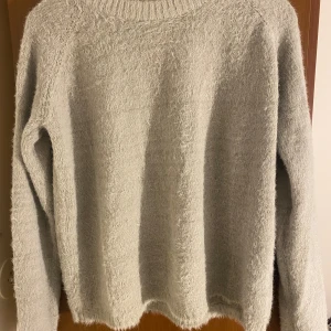 Ljusgrå fluffig stickad tröja M - Supermjuk och fluffig ljusgrå stickad tröja från The Black Label Collection i storlek M. Tröjan har långärmade ärmar och ribbad rund hals. Perfekt för dig som gillar en chill och cozy vibe under höst och vinter.