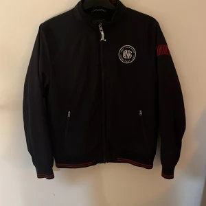 Mörkblå vindjacka från GANT - Snygg mörkblå vindjacka från GANT med dragkedja framtill och två fickor med zip. Jackan har röd broderad text på ärmen och ribbade muddar med röda ränder. Perfekt för dig som gillar sportig och clean stil. Jackan passar dig som har storlek xs, jackan är köpt på nk för cirka 1500 och ser dessutom helt ny - tun höstjacka 