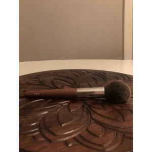 CAIA F16 sminkborste i brun - Sminkborste från CAIA, modell F16, med mjuka borst i gråbrunt och ett brunt handtag. Borsten har en rundad form och metallfäste i silver. Perfekt för applicering av puder, bronzer eller rouge. Snygg och stilren design som känns lyxig i handen.