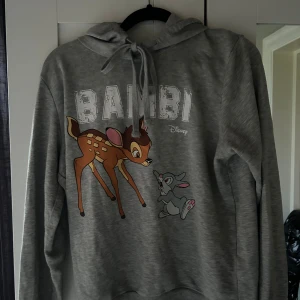 Grå Bambi hoodie  - Grå hoodie med Bambi-tryck från Disney ( new yorker) . Tröjan har snörning vid huvan, ribbade muddar och lång ärm. Mjuk bomullsmix som är perfekt för chill dagar. Stor Bambi och Stampe-tryck framtill ger en gullig vibe.