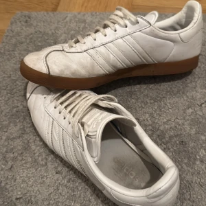 Adidas Gazelle vita sneakers - Klassiska Adidas Gazelle sneakers i vitt skinn med bruna gummisulor. Skorna har de ikoniska tre ränderna på sidorna och guldfärgad Gazelle-text. Lågt skaft och snörning framtill. Perfekta för en clean och sportig look. OBS de är 45 men passar även storlek 44 