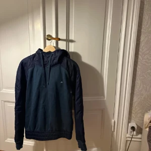 Mörkblå hoodie från J.Lindeberg - Snygg mörkblå hoodie från J.Lindeberg med huva och snörning. Tröjan har kontrasterande material på ärmar och axlar, samt ett litet broderat logomärke på bröstet. Perfekt för dig som gillar stilren och sportig look.