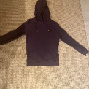 Mörklila hoodie från Lyle & Scott - Snygg mörklila hoodie från Lyle & Scott med klassisk gul logga på bröstet. Tröjan har huva med snörning, magficka och långa ärmar. Perfekt för chill dagar och enkel att matcha med jeans eller joggers.
