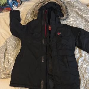 Svart parkajacka från Fjällräven - Svart parkajacka från Fjällräven med stor huva och snygg fuskpäls runt kanten. Jackan har flera praktiska fickor framtill, röd logotyp på bröstet och dragkedja med knappslå. Insidan är vadderad för extra värme och huvan har snörning för att justera passformen.