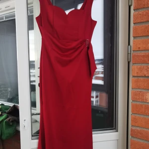 Röd långklänning med drapering - Oanvänd snygg röd långklänning i figurnära modell med breda axelband och hjärtformad urringning. Klänningen har draperad detalj i midjan med dekorativa stenar.