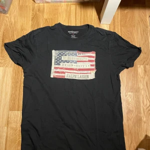 Svart t-shirt Ralph Lauren S - Svart t-shirt från Ralph Lauren Denim & Supply i storlek S. Tryck med amerikansk flagga och text på bröstet. Skön passform och tillverkad i 100% bomull. Perfekt för dig som gillar streetstyle och klassiska märken.