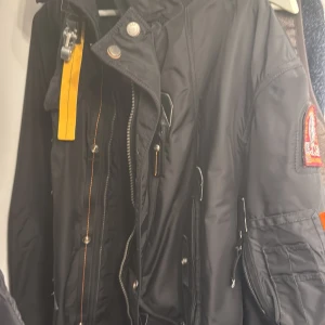 Svart Parajumpers dunjacka med äkta räv päls - Svart dunjacka från Parajumpers med ikonisk patch på ärmen, gul detalj vid halsen och päls på huvan. Jackan har flera fickor, dragkedja och knappar framtill samt orange detaljer. Perfekt för kalla dagar och har en cool, urban vibe. Den är äkta har kvitto köpte den för 12 tusen kronor allt är nytt och ej använd 